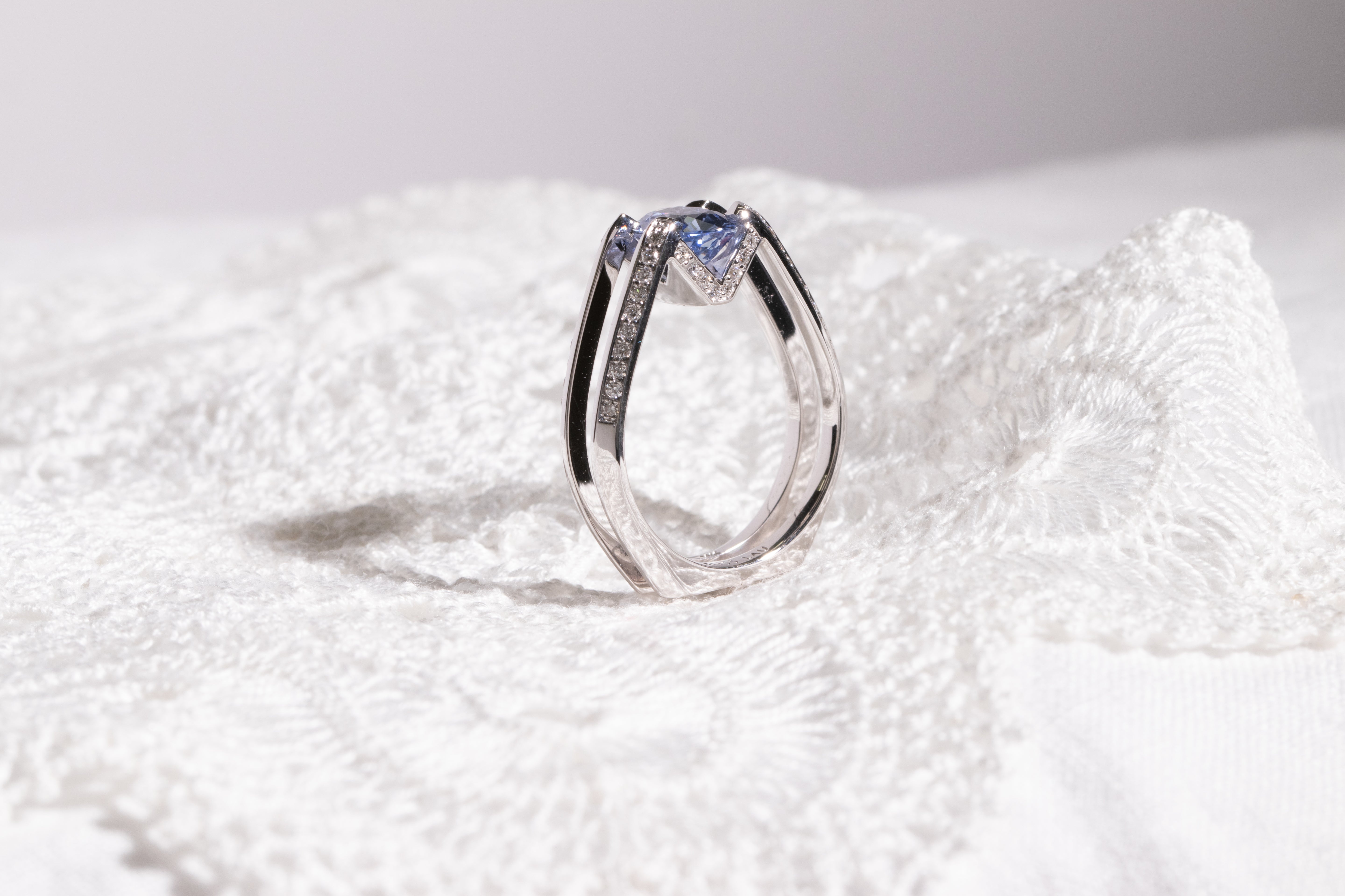 Futurist Engagement Ring Signature Collection highlighting an exquisite blue gemstone, elegantly perched on a delicate silver double band against a soft lace backdrop.