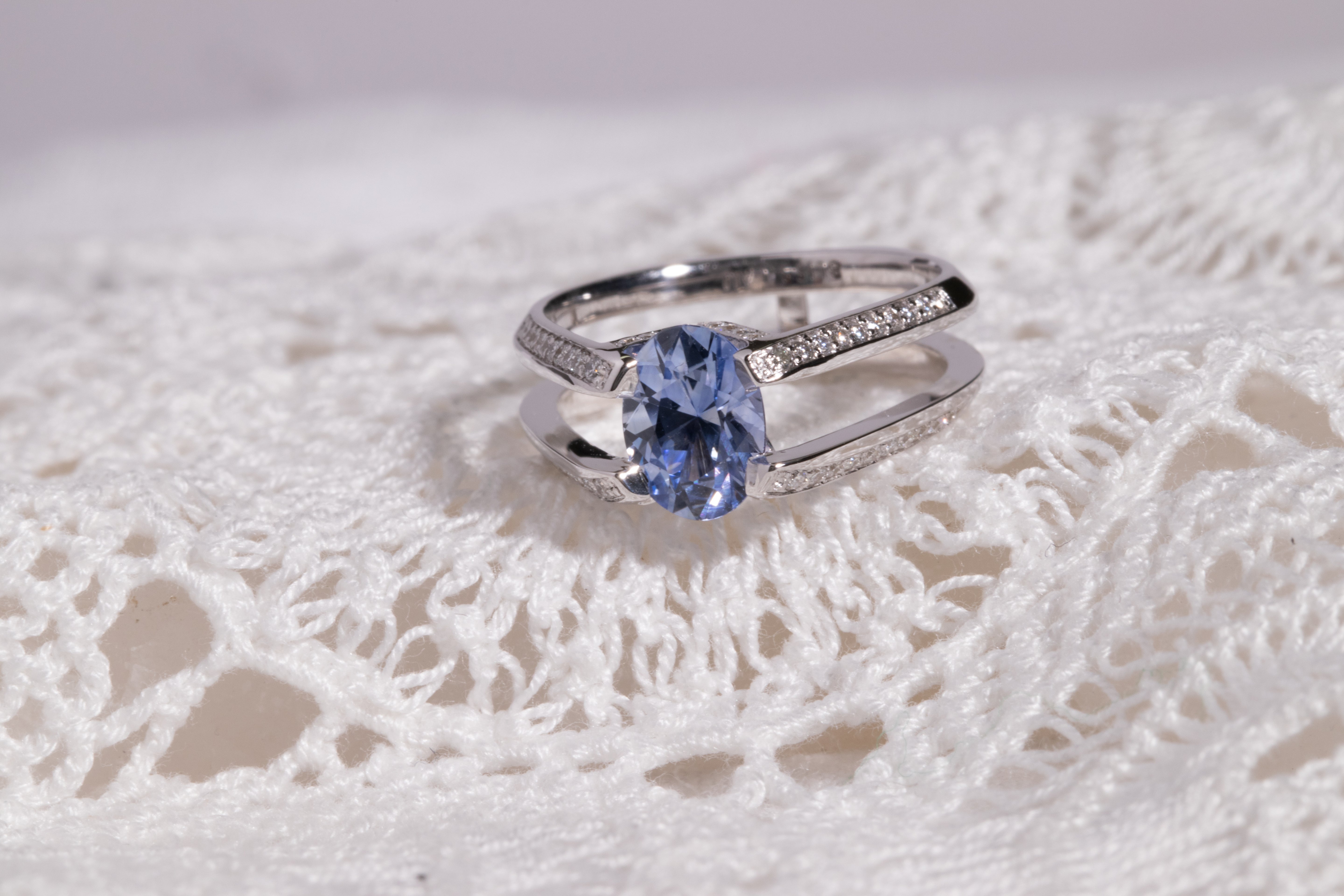 Futurist Engagement Ring Signature Collection showcasing a stunning oval blue gemstone, beautifully complemented by pave set diamonds on a polished silver band, resting on intricate lace.