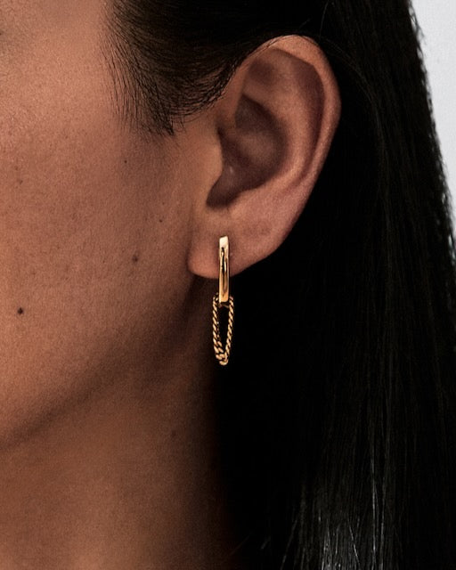 A close-up of a person's ear wearing a delicate Bliss Lau gold hoop earring with a twisted chain dangle, set against dark hair and skin tones.