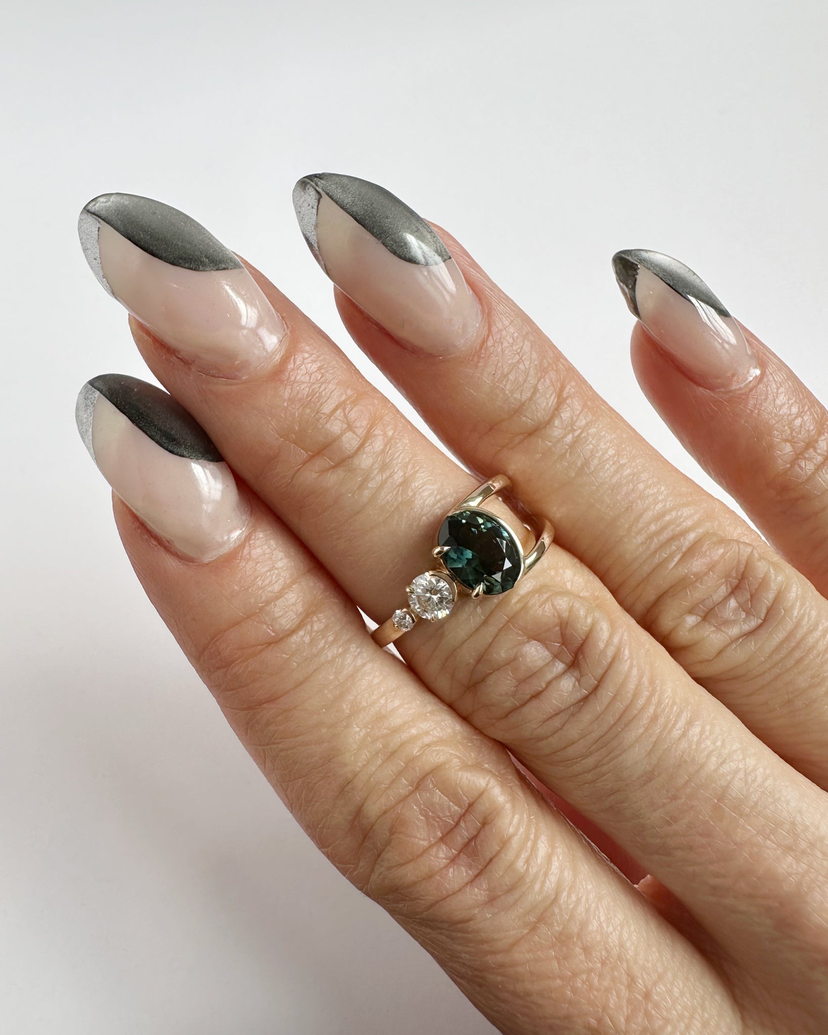 Close-up of a hand adorned with the Bright Ring Green Sapphire, featuring delicate nails with a stylish gray tip, showcasing luxury craftsmanship and a deep blue-green Australian sapphire centerpiece.