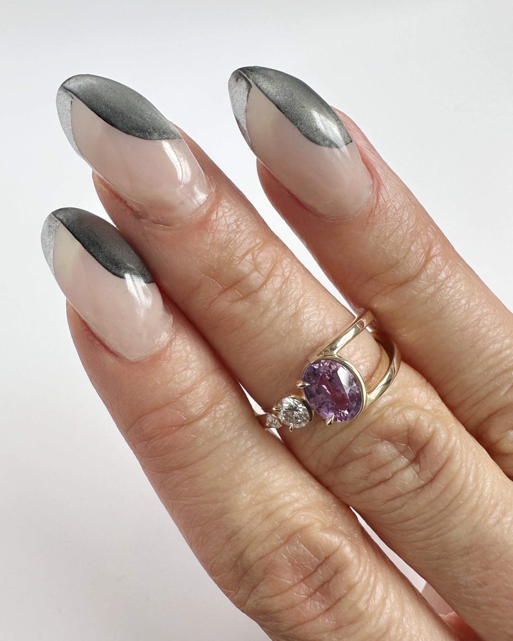 A close-up of a beautifully manicured hand showcasing the Bright Ring Lilac Sapphire, featuring a stunning oval pink lilac heritage sapphire set in 14K gold, adorned with a sparkling lab-created diamond. The elegant design complements the chic nail art, highlighting the ring's chic luxury aesthetic.