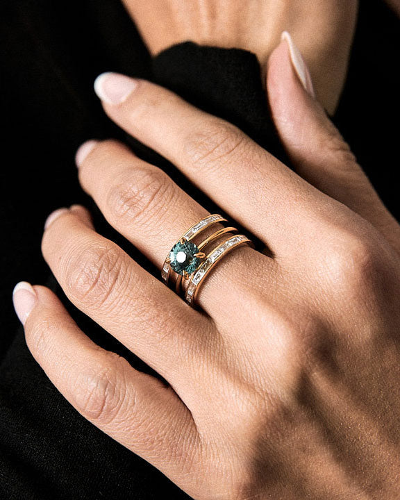 Hand wearing a gold ring set with a blue sapphire gemstone and diamond baguettes on a black background