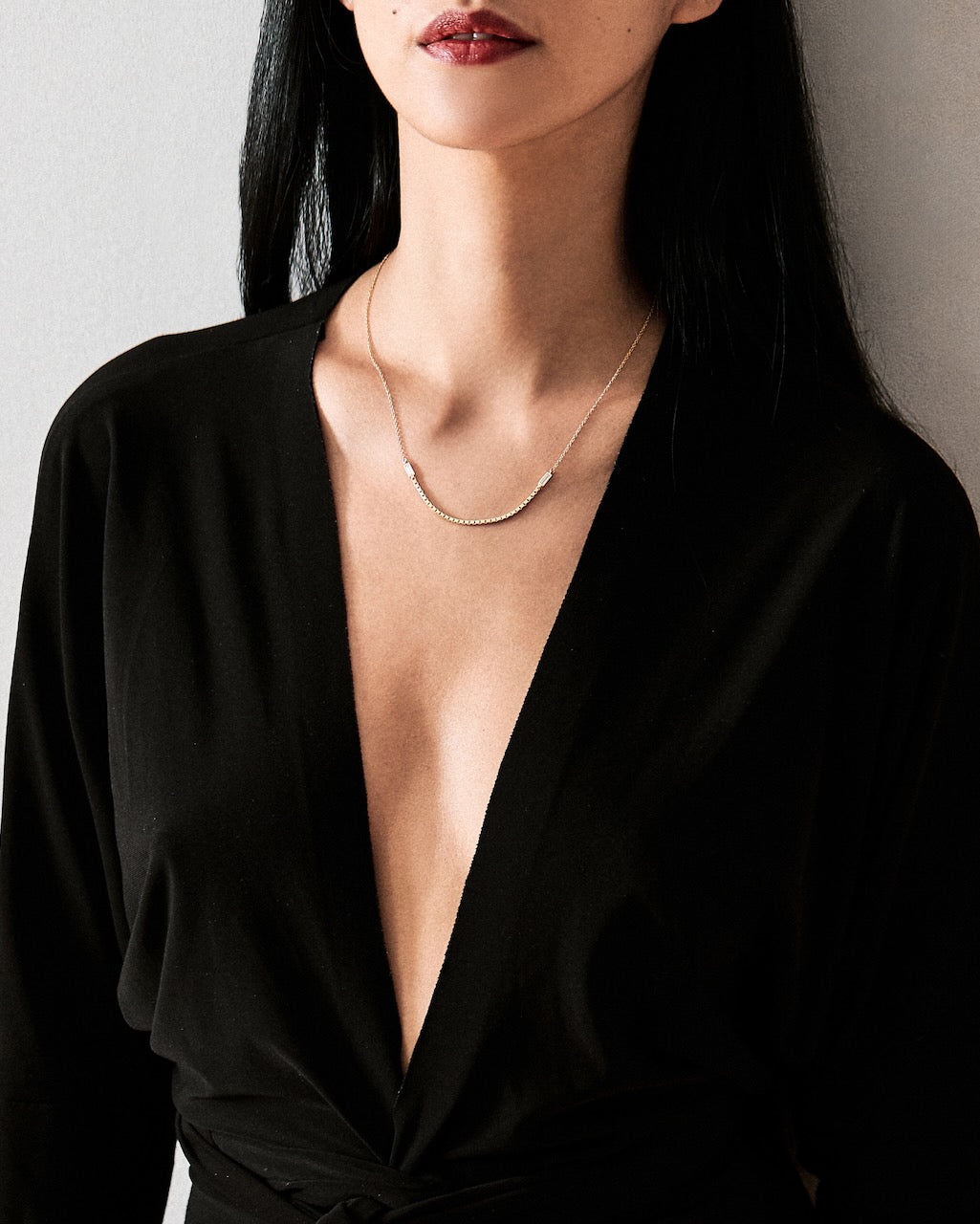 Person wearing a black garment with a deep neckline and a gold necklace against a neutral background