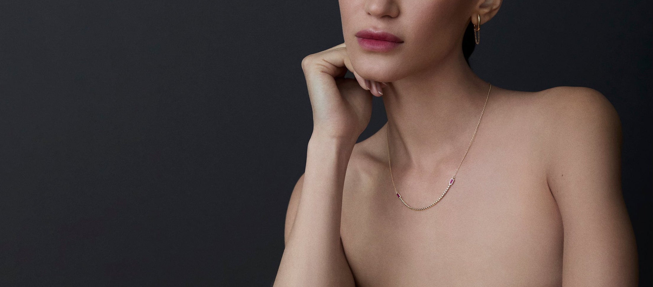 Woman wearing a delicate 18k gold and ruby necklace against a dark background
