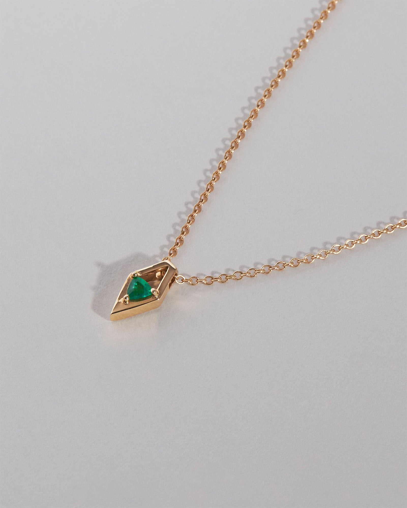 A delicate yellow gold chain necklace featuring a small geometric pendant with a vibrant emerald gemstone, photographed on a clean light gray background.
