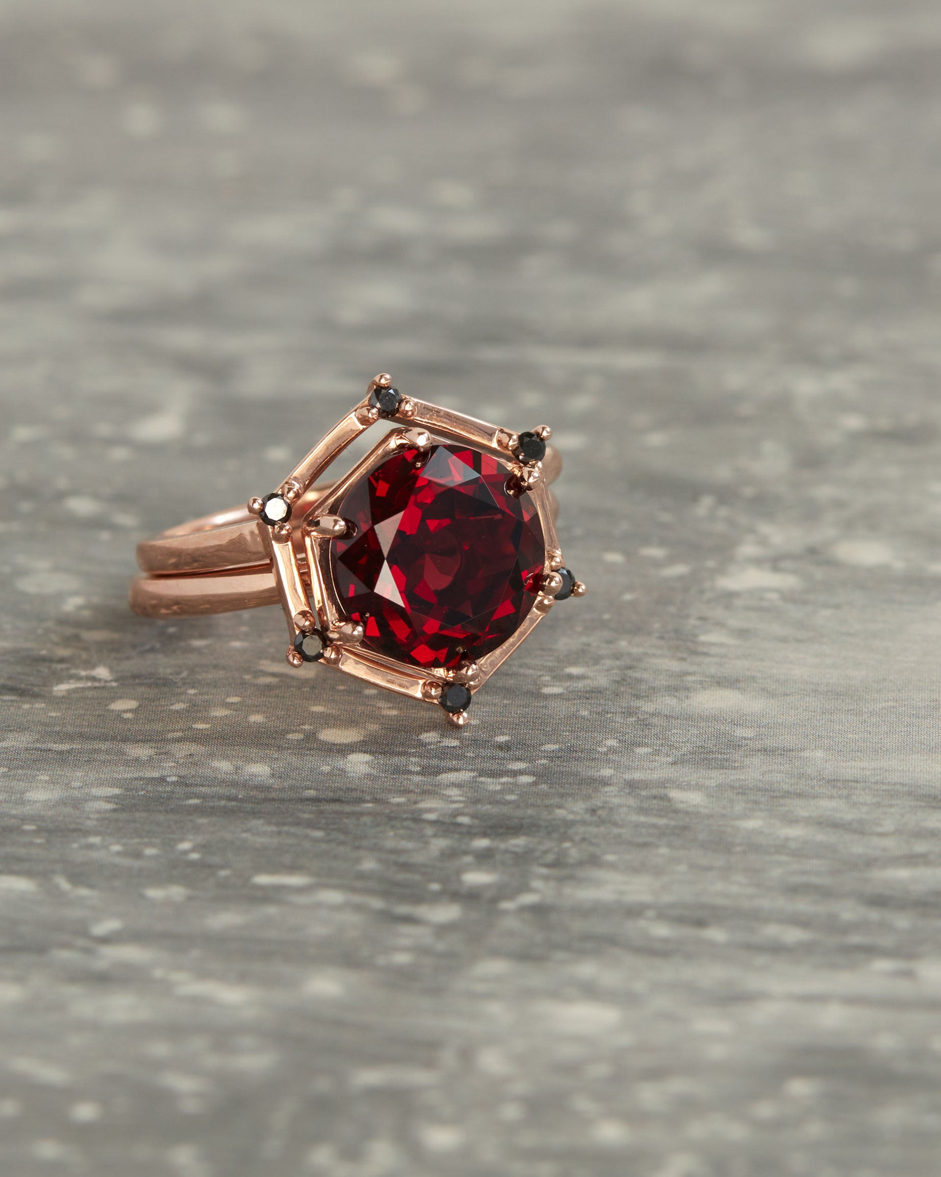 Aura Ring Wedding Ring Set elegantly posed on a textured surface, featuring a gorgeous round red garnet framed by sleek recycled gold and black diamonds.
