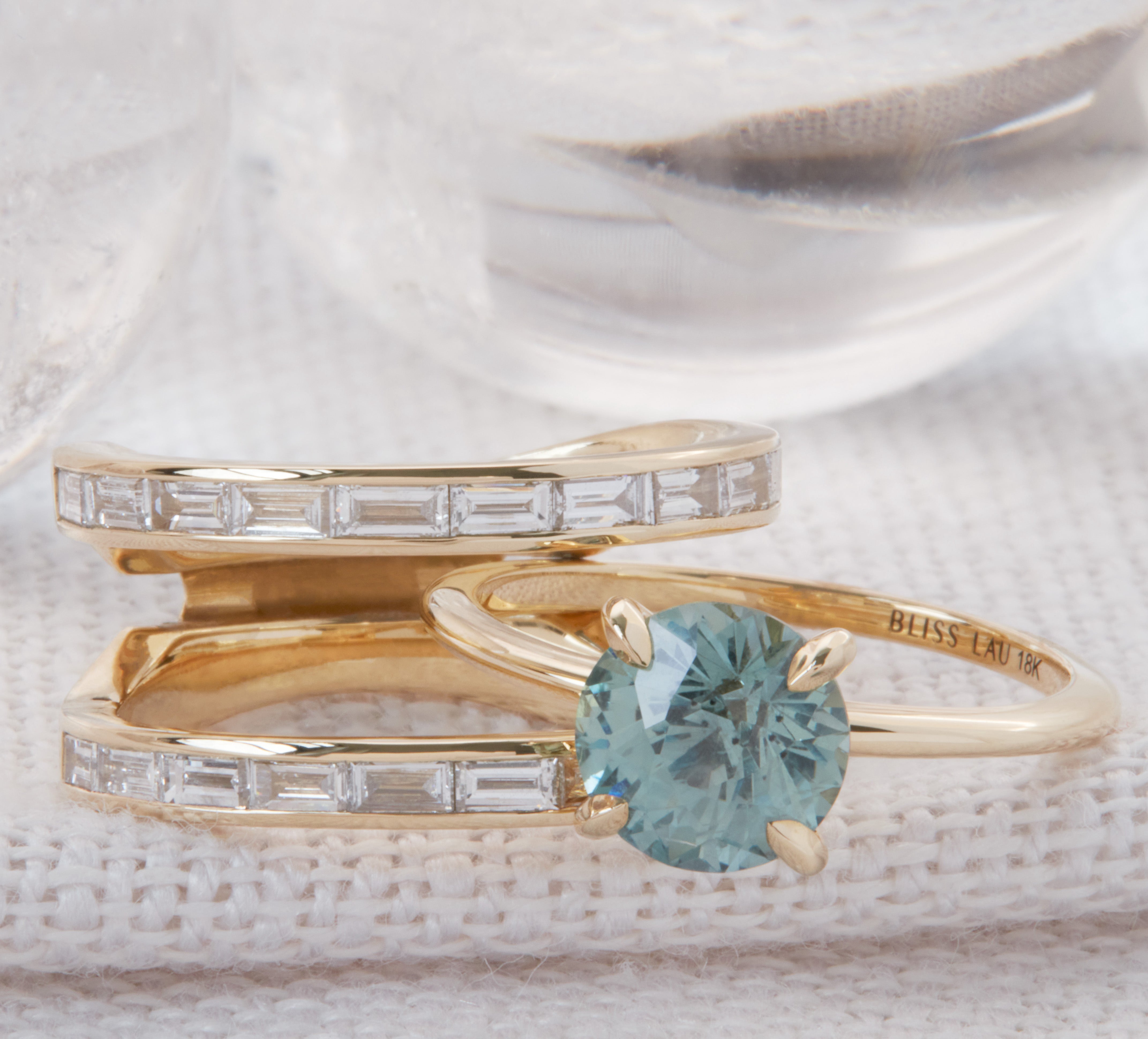 Cascade II Enclose Ring featuring a unique interlocking design with a stunning aqua gemstone surrounded by channel-set diamond baguettes, elegantly displayed against a soft textured background.