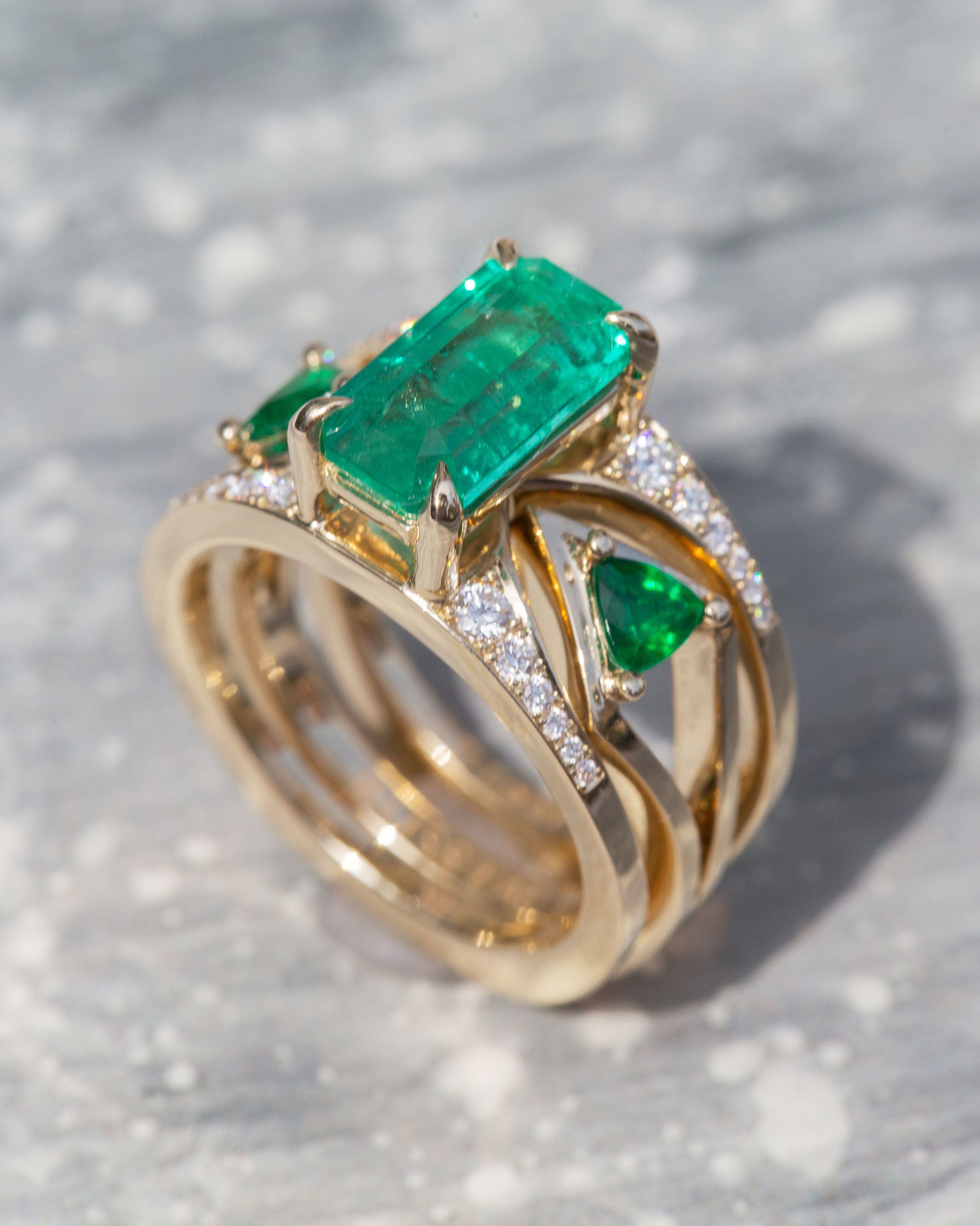 Artistic shot of the Mirage + Reflection Ring Set Emerald on a textured surface, emphasizing the brilliance of the emeralds and elegant gold craftsmanship.