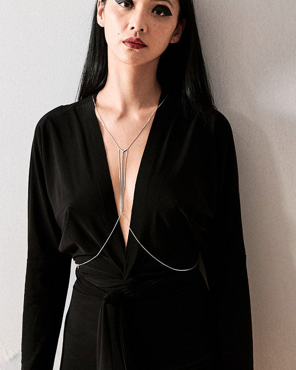 A model wearing a minimalistic body chain over a light grey top, showcasing the elegant design against a simple backdrop. The bodychain adds a stylish touch to her outfit, emphasizing the intricate details of the jewelry.