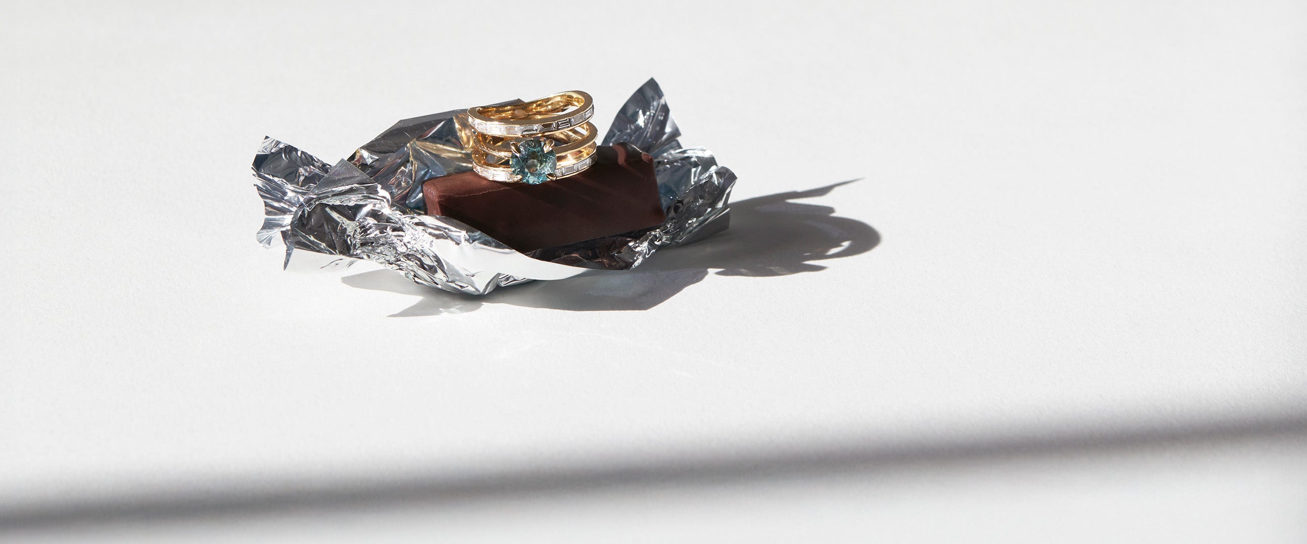 gold and sapphire ring set with baguette diamonds one piece of chocolate and crumpled wrapper on a white background