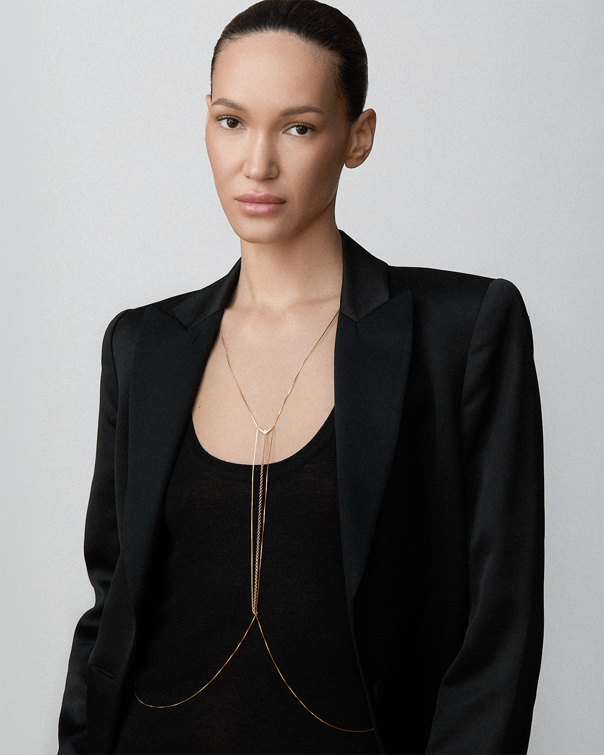 A model wearing a Bliss Lau gold Interlude body chain over a black top and blazer, set against a plain light gray background.