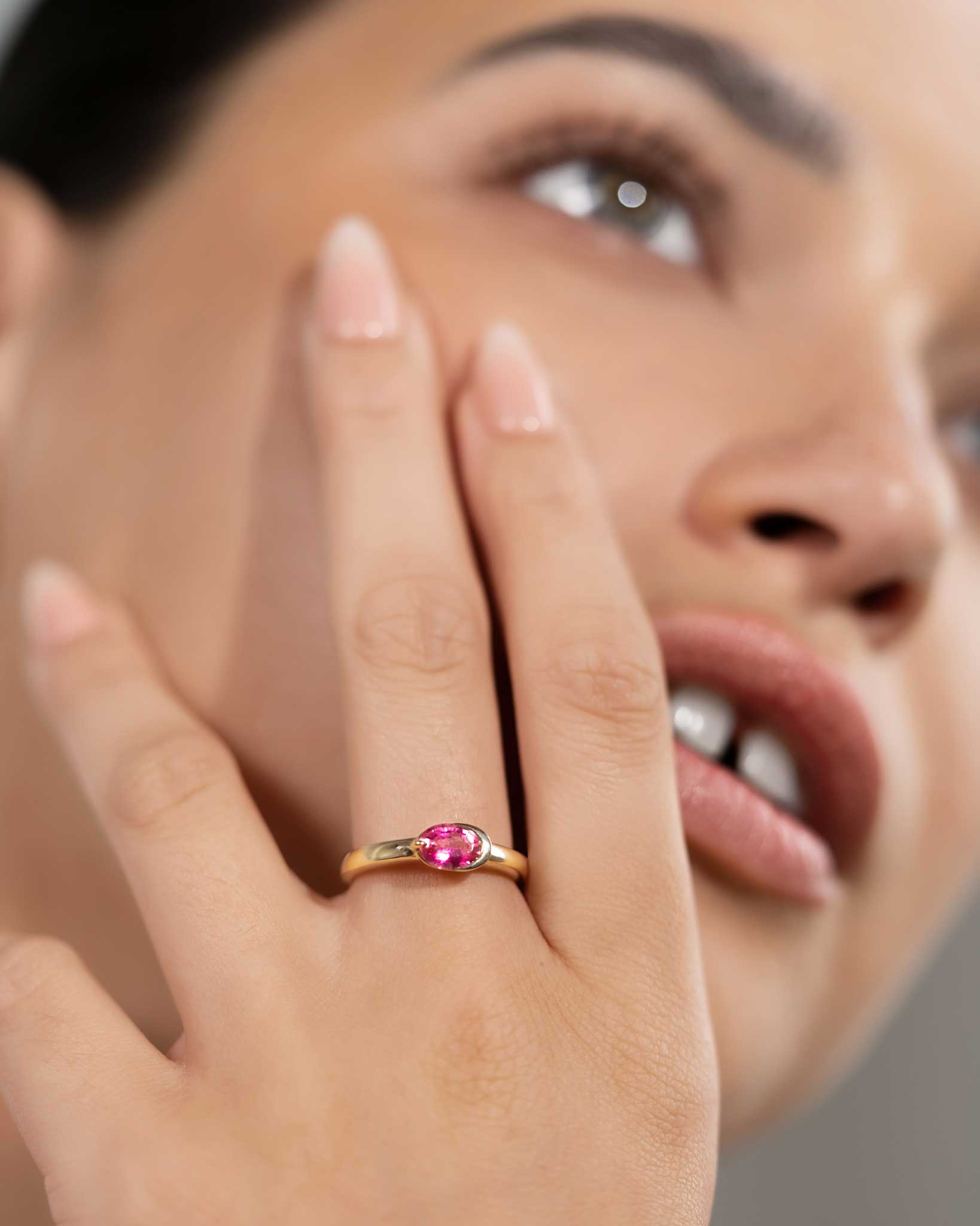 Close-up of a hand wearing a gold ring with a pink gemstone, held near the face.