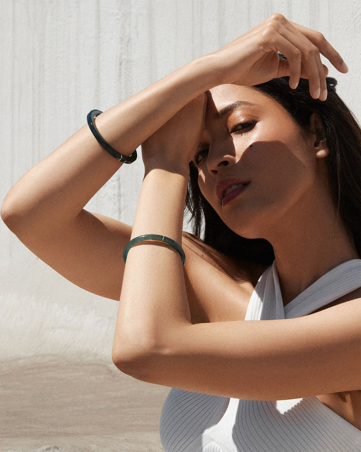 A woman wearing a Bliss Lau  jade bracelet on either wrist, posing against a textured light gray concrete wall.